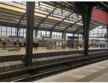 Großer Bahnhof mit Dachkonstruktion aus Glas und Stahl, darunter Bahnsteige und Gleise. Im Bild ist der Bahnhof Berlin Friedrichstraße zu erkennen, mit Menschen auf den Bahnsteigen und einem vorbeifahrenden ICE-Zug.