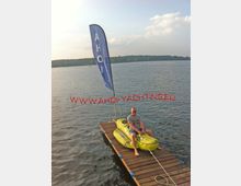 Mann sitzt auf einem gelben, krokodilförmigen Schlauchboot, das auf einem kleinen hölzernen Steg auf einem ruhigen See befestigt ist. Im Hintergrund ist eine blaue Fahne mit der Aufschrift „AHOI“ sowie bewaldetes Ufer zu sehen.