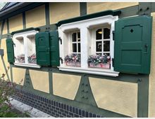 Gelbes Fachwerkhaus mit dunkelgrünen Holzbalken und grünen Fensterläden, die teilweise geöffnet sind. Vor den Fenstern stehen Blumenkästen mit bunten Blumen, darunter eine Mauer mit dunklen Ziegeln und hell gepflastertem Weg.