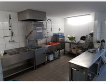 Industrieküche mit Edelstahlmöbeln, einschließlich einer Spüle mit zwei Becken, einer großen Geschirrspülmaschine, Arbeitstischen und Regalen. Die Wände sind mit weißen Fliesen verkleidet, und das Beleuchtungssystem ist in die Decke integriert.