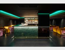 Moderne Bar mit zentraler, grüner Marmor-Theke, umgeben von dimmer Beleuchtung. Im Hintergrund beleuchtete Regale mit Flaschen und Gläsern; Sitzbereiche mit samtigen gelben und lilafarbenen Sesseln befinden sich an den Seiten.