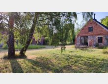 Ein weitläufiger Garten mit Bäumen und Rasen, im Hintergrund ein rustikales Gebäude aus roten Ziegeln und Natursteinen. Im Vordergrund stehen Holzbänke und ein Tisch auf einer grasbewachsenen Fläche.