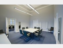 Konferenzraum mit einem quadratischen Tisch, umgeben von acht Polsterstühlen mit Holzarmlehnen. Der Raum ist hell, mit grauen Wänden, blauem Teppichboden und schlichten Deckenleuchten; im Hintergrund ist ein weiterer Raum mit Fenstern und Konferenztischen zu sehen.