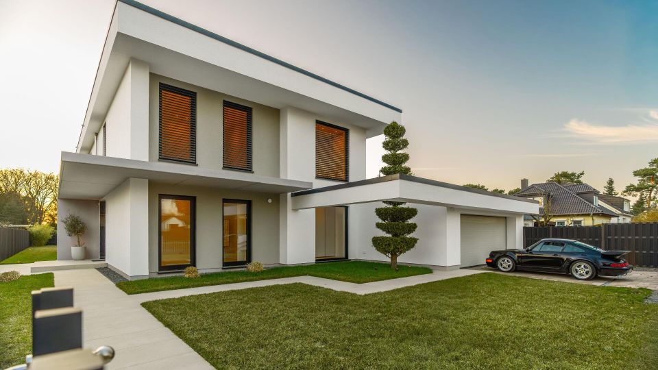 Modernes, zweigeschossiges Wohnhaus mit flachem Pultdach, weiß-grauer Fassade und großen Fenstern mit Lamellenjalousien. Im Vordergrund liegen ein gepflegter Rasen, ein gepflasterter Weg und eine angrenzende Garage mit Einfahrt, eingefasst von einem dunklen Zaun in einer Wohnsiedlung.