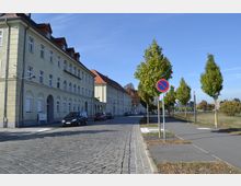 Straßenszene mit gepflastertem Fahrweg und einem gelblichen historischen Wohngebäude auf der linken Seite, das durch Bögen und viele Fenster gekennzeichnet ist. Rechts führt ein Gehweg mit Bäumen entlang einer Wiese, mehrere Autos sind am Straßenrand geparkt, und ein Parkverbotsschild ist sichtbar.