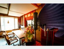 Ein Aufnahmestudio mit einem Mikrofon im Vordergrund, umgeben von einer schallabsorbierenden Halterung. Im Hintergrund befinden sich ein Schreibtisch mit Bürostühlen, ein Bildschirm, Pflanzen sowie eine Fensterfront, die Tageslicht hereinlässt.