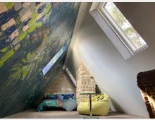 Dachgeschossraum mit schrägen Wänden, darunter eine Wand mit farbenfrohem, abstraktem Muster. Ein Dachfenster lässt Tageslicht herein, und im Raum befinden sich eine niedrige Schlafnische, ein gelber Sitzsack, ein kleiner Glastisch und eine gemauerte Backsteinwand.