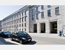 Straßenansicht eines großen, mehrstöckigen Gebäudes aus grauem Stein mit rechteckigen Fenstern in symmetrischer Anordnung, breiten Eingangstoren und einem gepflasterten Bürgersteig. Im Vordergrund sind parkende Autos und gehende Passanten sichtbar.