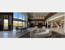 Großer, eleganter Wohnzimmerbereich mit hohen Decken, verziert mit Holzvertäfelung und einem großen Kronleuchter. Der Raum umfasst ein Sofa mit Kissen, einen Esstisch mit Stühlen vor einer Glasfront mit Aussicht auf eine grüne Landschaft, sowie eine verzierte Treppe und ein Geländer im Hintergrund.