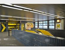 Innenansicht einer U-Bahn-Station mit blauen Wandfliesen, gelben Rolltreppen und einem Kiosk ("Backshop") im Hintergrund. Eine Uhr und ein Fahrkartenautomat sind an der Wand angebracht, während große Fenster auf der rechten Seite Tageslicht hereinlassen.