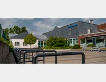 Außenansicht eines Gebäudekomplexes mit der Beschriftung „Schwimmhalle“ auf einer grauen Metallfassade. Im Vordergrund sind Fahrradständer und ein gepflasterter Weg, daneben grüne Büsche, ein Baum und Blumenbeete vor großen Fenstern.