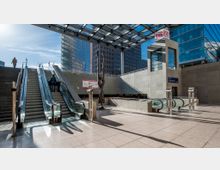 Eingangsbereich eines modernen Bahnhofs mit Rolltreppen und gläsernem Dach. Im Hintergrund sind Bürogebäude, eine abstrakte Skulptur aus Metall und ein DB-Logo zu sehen.