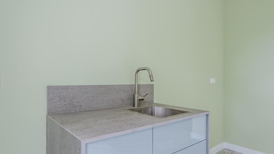 Moderne Küchenzeile mit grauer Beton-Arbeitsplatte und -Rückwand, integriertem Edelstahlspülbecken und hohem Wasserhahn. Glatte, hellgraue Fronten stehen vor einer hellgrünen Wand in einem schlichten Innenraum.