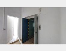 Weiße Innenraumwand mit einer offenen Metalltür, die zu einem kleinen Lastenaufzug führt. Rechts neben der Tür befindet sich ein Aufzug-Bedienfeld, und links ist ein heller, gefliester Durchgang mit einer Tür sichtbar.