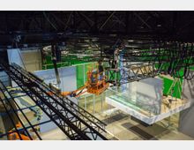 Technische Produktionshalle mit einem Arbeiter auf einer Hebebühne, der an einer Metallträgerkonstruktion unter der Decke arbeitet. Im Hintergrund sind grüne Wände, Beleuchtungselemente und weiße Stellwände sichtbar.