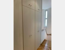 Schmaler Flur oder Treppenhausbereich mit einer langen Reihe hoher, weißer Einbauschränke entlang der linken Wand. Rechts führt eine Holztreppe mit Geländer nach unten, daneben ein schmales, hohes Fenster, das Tageslicht in den Raum lässt.