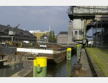 Industriehafen mit Anlegestellen und mehreren rostigen Frachtschiffen, flankiert von alten Lagerhäusern und gelben Pollern entlang des Kais. Im Hintergrund sind ein Backsteingebäude mit Turm sowie moderne Bürogebäude zu sehen.