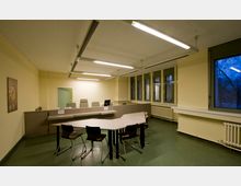 Büroraum mit gelb gestrichenen Wänden, grünem Bodenbelag und mehreren rechteckigen Tischen und Stühlen in der Mitte. Große Fenster mit grünem Rahmen befinden sich an der rechten Wand, darunter Heizkörper, und eine Tür ist im Hintergrund sichtbar.