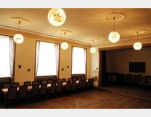 Ein Versammlungsraum mit langen, aneinandergereihten Tischen und gepolsterten Stühlen. Der Raum hat helle Wände, mehrere große Fenster mit halbtransparenten Vorhängen, hölzernen Parkettboden und auffällige hängende Kugelleuchten an der Decke.