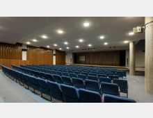 Ein moderner Vortragssaal mit mehreren Reihen blauer gepolsterter Sitzplätze, einer Bühne im Hintergrund, die von einem Vorhang umrahmt ist, und einer Projektionsfläche in der Mitte. Die Wände sind teilweise mit Holzverkleidungen gestaltet, und die Decke ist mit gleichmäßig angeordneten Deckenleuchten beleuchtet.