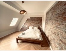 Dachgeschoss-Schlafzimmer mit schräger Decke, unverputzten Ziegelwänden und einem Holzbett mit weißer Bettwäsche. Ein Dachfenster und ein hölzerner Fischgrät-Parkettboden prägen den Raum, der durch eine kupferfarbene Hängeleuchte ergänzt wird.