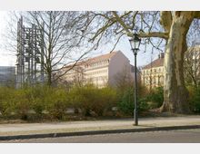 Ein städtischer Park mit Gehweg, Laternenmast und großen Bäumen im Vordergrund. Im Hintergrund sind ein modernes Glockengestell und mehrere historische Gebäude mit roten Ziegeldächern sichtbar.