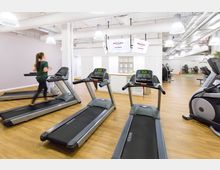 Innenraum eines modernen Fitnessstudios mit hellen Wänden und Holzboden. Im Vordergrund befinden sich mehrere Laufbänder, eines wird von einer Person verwendet, während im Hintergrund andere Fitnessgeräte und eine Sitzbank sichtbar sind.