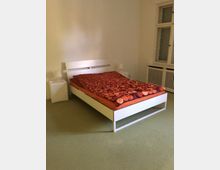 Schlafzimmer mit einem weißen Bettgestell und einer orange-rot gemusterten Bettdecke, flankiert von zwei weißen Nachttischen mit kleinen Lampen. Im Hintergrund ist eine helle Wand, ein Fenster mit Fensterbrett und ein Heizkörper zu sehen.