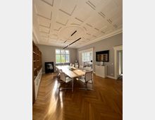 Helles Konferenzzimmer mit einem langen Holztisch, umgeben von weißen Stühlen auf einem Parkettboden im Fischgrätmuster. Die Wände sind grau, mit großen Fenstern, einer dekorativen Decke, einem eingebauten Regal sowie einem Flachbildschirm an der Wand.