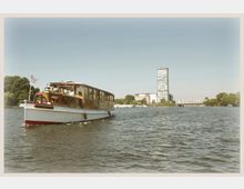 Ein weißes Ausflugsboot mit hölzernem Aufbau fährt auf einem Fluss. Im Hintergrund sind moderne Bürogebäude, eine Brücke sowie Bäume an den Ufern zu sehen.