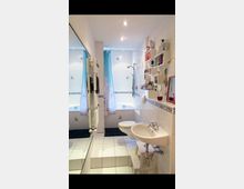 Helles Badezimmer mit weiß gefliesten Wänden und Böden, ausgestattet mit einer Badewanne, einem weißen Waschbecken, einer Toilette und einem Handtuchheizkörper. An der rechten Wand befinden sich offene Regale mit Pflegeprodukten, und ein großer Spiegel bedeckt die linke Wand.