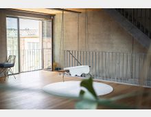 Loftartiger Innenraum mit Sichtbetonwänden, Holzdecke und Holzfußboden, in dem eine hängende Wiege über einem runden Teppich befestigt ist. Links führt eine große Glas-Schiebetür auf einen Balkon, rechts verläuft eine Metalltreppe entlang der Wand.