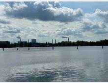 Seeufer mit Blick auf ein Gewässer, im Hintergrund eine Industrieanlage mit rauchenden Schornsteinen und einem großen Kühlturm sowie Bäume entlang des Ufers. Der Himmel ist bewölkt, und die Wasserfläche wird von mehreren Markierungen durchzogen.