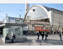 U-Bahnhof Alexanderplatz in Berlin mit einem modernen gläsernen Eingangsbereich im Vordergrund und dem markanten Schriftzug „Alexanderplatz“ auf einem Gebäude mit gewölbter Glasfassade im Hintergrund. Menschen laufen und sitzen auf dem Platz, während Straßenbahnschienen durch die Szene verlaufen.