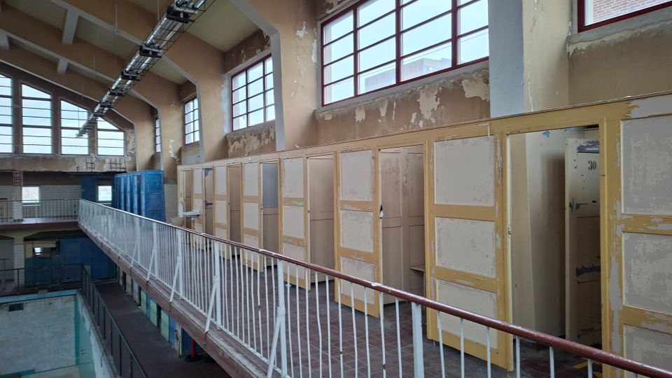 Ehemaliger Innenraum eines Schwimmbads mit Umkleidekabinen aus Holz, abgenutzten gelb-beigen Türen und abblätternder Farbe. Oberlichter mit roten Fensterrahmen lassen Tageslicht herein, und eine Metallbrüstung grenzt die obere Etage vom leeren Schwimmbecken ab.