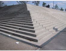 Große, breite Freitreppe aus Beton mit klaren geometrischen Linien; umgeben von einer gepflasterten Fläche mit vereinzeltem Müll auf dem Boden. Im Hintergrund sind Bäume und ein paar Personen erkennbar, bei sonnigem Wetter.