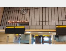 Innenansicht eines Flughafenterminals mit einer großen Wanduhr, neonfarbenem Schriftzug „Restaurant“ und mehreren gelben Abfluganzeigetafeln. Im Hintergrund Glas- und Metallstrukturen sowie ein Durchgangsbereich.