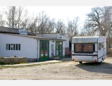 Ein kleiner weißer Gebäudekomplex mit grünen Türen und einem Schild mit der Aufschrift „fabrik“ neben einem geparkten, leicht verwitterten Wohnwagen auf einem unbefestigten Gelände, umgeben von kahlen Bäumen.