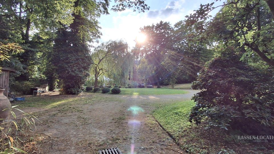 Weitläufiger Park mit breitem, unbefestigtem Weg durch Rasenflächen, umgeben von hohen Laubbäumen, Sträuchern und einer Weide im Hintergrund. Links stehen ein hölzerner Schuppen und große Tonkrüge, während die tief stehende Sonne durch das Blätterdach scheint.