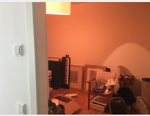 Ein unaufgeräumtes Zimmer mit warmem, orangefarbenem Licht. Im Raum befinden sich ein Synthesizer an der Wand, gestapelte Bilderrahmen, ein Stuhl mit abgelegten Kleidungsstücken und diversen Gegenständen sowie Kartons und kleinere Möbelstücke auf Holzboden.