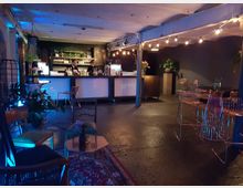 Eine stilvolle Bar in einem dunklen, modernen Raum mit einer langen Theke, hinter der zwei Personen stehen, und Regalen mit Gläsern im Hintergrund. Der Raum ist mit Stehtischen, Stühlen, Pflanzen und dezentem Licht sowie dekorativen Glühbirnen gestaltet.