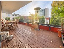 Großzügige Terrasse mit Holzdielen, ausgestattet mit Holzmöbeln, Heizstrahler und mehreren großen Topfpflanzen, darunter Palmen; im Hintergrund sind Wohnhäuser, Bäume und ein moderner Gebäudekomplex zu sehen.