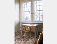 Ein schlichtes Zimmer mit einem Holzschreibtisch und passendem Stuhl mit Metallgestell auf einem bunt gemusterten Teppich. Der Schreibtisch steht vor einem großen Fenster mit weißem Rahmen, durch das Bäume und helles Tageslicht sichtbar sind, im Hintergrund befindet sich ein Heizkörper.