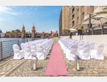 Außenbereich einer Hochzeitszeremonie mit Stühlen, die in zwei Reihen auf Kopfsteinpflaster aufgestellt sind, getrennt durch einen rosa Teppich. Im Hintergrund ist die Oberbaumbrücke über die Spree in Berlin zu sehen, daneben ein Gebäude mit einer Terrasse.