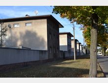 Reihe von Mehrfamilienhäusern mit schlichter Fassade und Flachdächern entlang einer Straße, die von Bäumen gesäumt ist. Im Vordergrund verläuft eine Mauer, und auf den Dächern sind Satellitenschüsseln sichtbar.