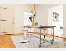 Heller Raum mit Holzboden, einem Tisch aus Metallrohren auf Rollen, umgeben von weißen Hockern, und einer Vase mit gelben Ziergräsern. Im Hintergrund ein angrenzender Raum mit Fotostudio-Ausrüstung und einer Wanddekoration aus bunten Scheiben.