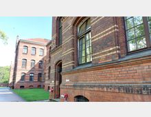 Backsteingebäude mit großen Fenstern und gelben Klinkerstreifen, umgeben von einer grünen Rasenfläche und einem gepflasterten Weg. Im Hintergrund stehen weitere Gebäude aus rotem Backstein, eingebettet in eine parkähnliche Umgebung.