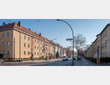 Straßenansicht mit beige- und pastellfarbenen Mehrfamilienhäusern mit roten Ziegeldächern, die entlang einer asphaltierten Straße stehen. Am Straßenrand sind karge Bäume, ein Fußweg sowie Verkehrsschilder und eine Straßenlaterne zu sehen; ein Auto fährt in der rechten Fahrspur.