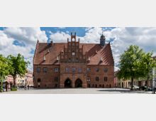 Backsteingebäude im gotischen Stil mit steilem Ziegeldach, filigraner Giebelgestaltung und einem kleinen Turm mit Kuppel. Im Vordergrund ein gepflasterter Platz, umgeben von Bäumen und farbigen Wohnhäusern.