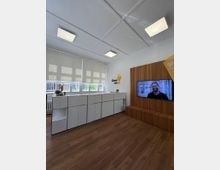 Ein moderner Büroraum mit Holzfußboden, weißen Wandschränken vor einem Fenster mit Rollos im Hintergrund, und einer Holzwand mit integriertem Bildschirm, auf dem ein Mann abgebildet ist. Die Raumdecke ist mit hellen quadratischen Deckenleuchten ausgestattet.